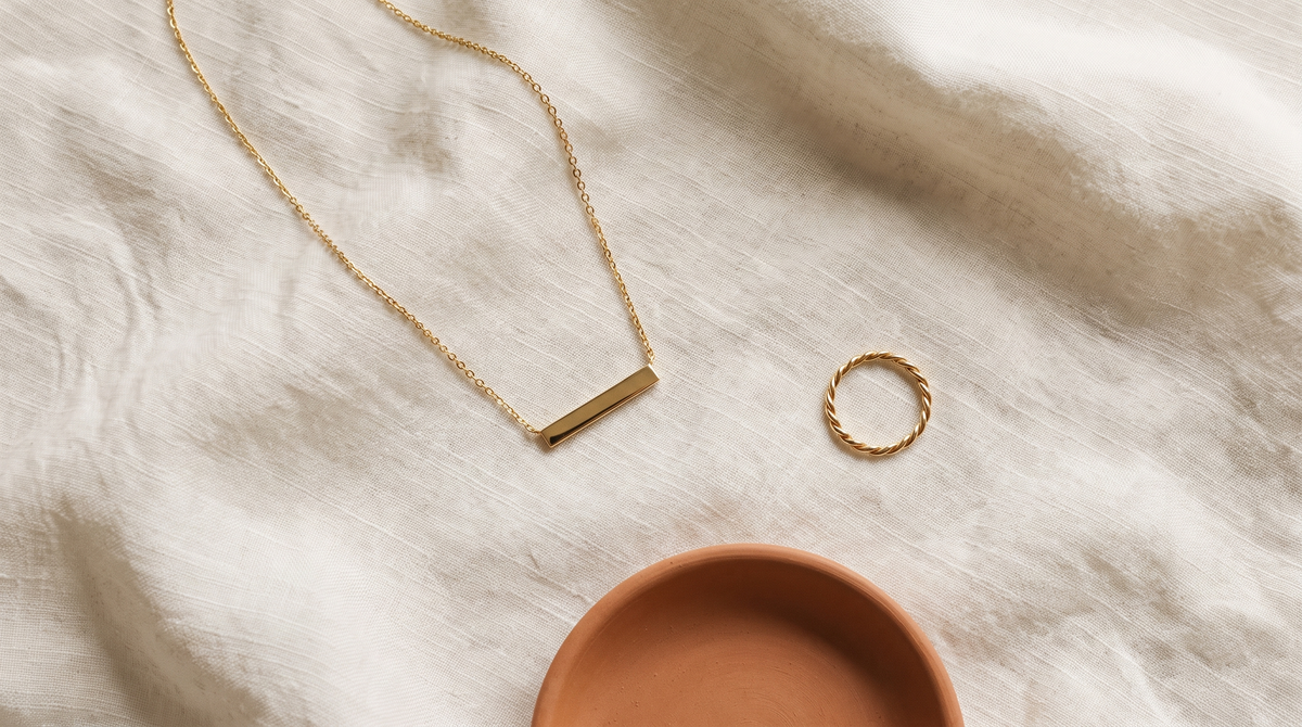 Gold bar necklace and twisted rope ring resting on ivory linen beside a ceramic dish