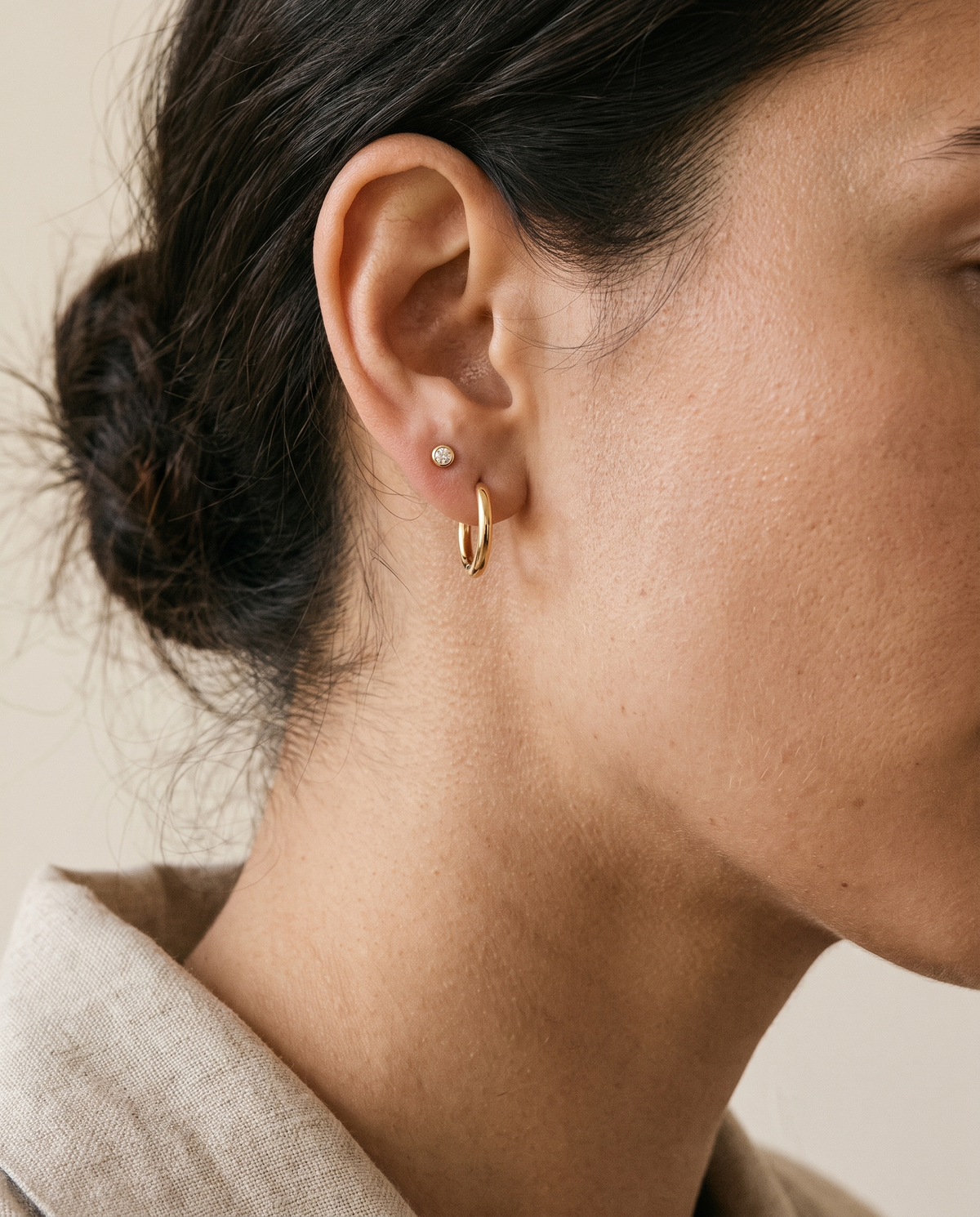 Ear wearing a small gold oval huggie with a bezel stud above it