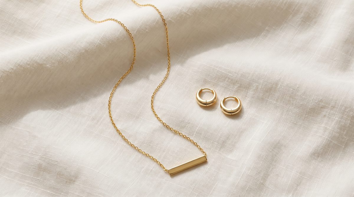Gold bar necklace and dome huggie earrings arranged with space between them on ivory linen